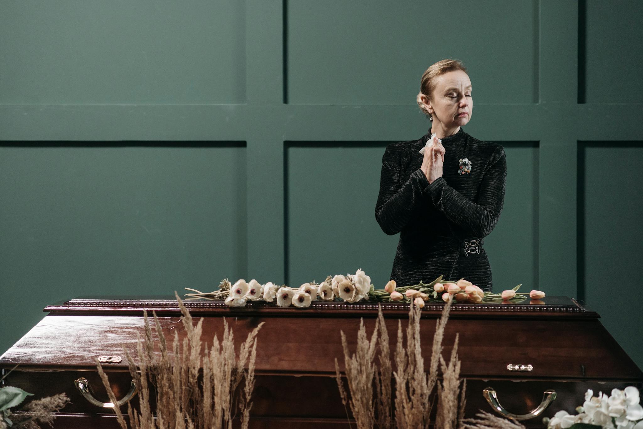 A solemn funeral scene with a woman mourning near a closed casket adorned with flowers.